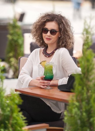 Foto de Chica morena feliz con gafas de sol sentada en el parque, bebiendo un vaso de jugo verde frío mientras sonríe a la distancia. Joven mujer bonita en el banco bebiendo jugo usando una camisa blanca - Imagen libre de derechos