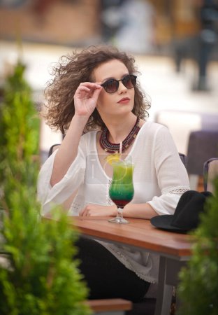 Photo for Happy Brunette Girl with sunglasses Sitting at the Park, Drinking a glass of Cold Green Juice While Smiling Into the Distance. Young pretty woman on the bench  drinking  juice wearing a white shirt - Royalty Free Image