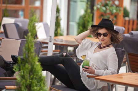 Foto de Chica morena feliz con gafas de sol y sombrero negro sentado en el parque, bebiendo un vaso de jugo verde frío Joven mujer bonita en el banco bebiendo jugo usando una camisa blanca - Imagen libre de derechos