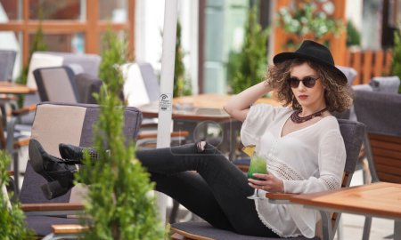 Foto de Chica morena feliz con gafas de sol y sombrero negro sentado en el parque, bebiendo un vaso de jugo verde frío Joven mujer bonita en el banco bebiendo jugo usando una camisa blanca - Imagen libre de derechos