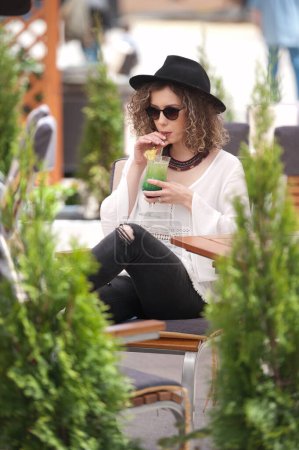 Téléchargez les photos : Joyeux Brunette Fille avec lunettes de soleil et chapeau noir Assis au parc, Boire un verre de jus vert froid Jeune jolie femme sur le banc boire du jus portant une chemise blanche - en image libre de droit