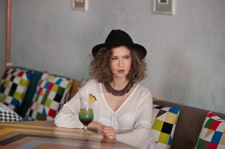 Foto de Happy Brunette Girl with black hat  Sitting indoor in bar, Drinking a glass of Cold Green Juice While Smiling Into the Distance. Young pretty woman on the bench  drinking  juice wearing a white shirt - Imagen libre de derechos