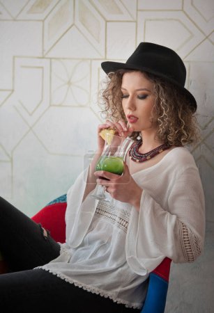 Photo for Happy Brunette Girl with black hat  Sitting indoor in bar, Drinking a glass of Cold Green Juice While Smiling Into the Distance. Young pretty woman on the bench  drinking  juice wearing a white shirt - Royalty Free Image