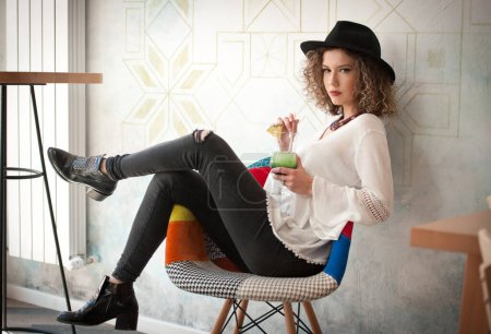 Téléchargez les photos : Happy Brunette Girl with black hat  Sitting indoor in bar, Drinking a glass of Cold Green Juice While Smiling Into the Distance. Young pretty woman on the bench  drinking  juice wearing a white shirt - en image libre de droit