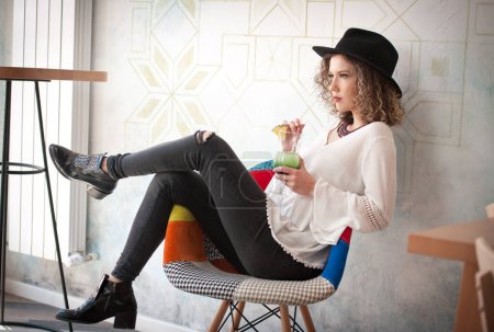 Foto de Happy Brunette Girl with black hat  Sitting indoor in bar, Drinking a glass of Cold Green Juice While Smiling Into the Distance. Young pretty woman on the bench  drinking  juice wearing a white shirt - Imagen libre de derechos