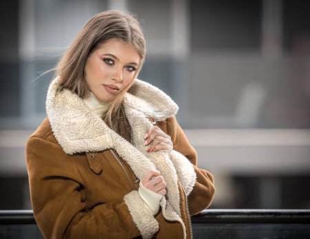Téléchargez les photos : Gorgeous smiling girl in stylish clothes posing on modern balcony with amazing view on city .Portrait of a Stylish fashionable blonde  teenage with fur coat with large collars wearing in balcony - en image libre de droit