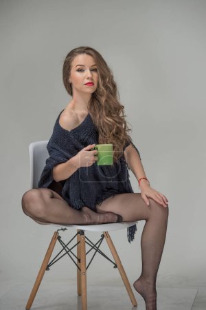 Photo for Attractive woman in black pantyhose and long legs posing challenging with a cup in hand. Classic boudoir scene. Indoor erotic photo. Sensual young  woman with long curly hair sit on the chair - Royalty Free Image
