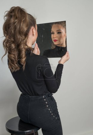 Téléchargez les photos : Belle femme en chemisier noir et lèvres rouges regarder dans le miroir devant un fond. Scène boudoir. Sensuelle jeune femme avec de longs cheveux bouclés et un jean noir devant la caméra - en image libre de droit