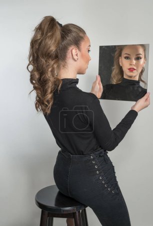 Photo for Attractive woman in black blouse and red lips  look into the mirror in front of a background. boudoir scene.  Sensual young  woman with long curly hair and black jeans wearing in front of camera - Royalty Free Image