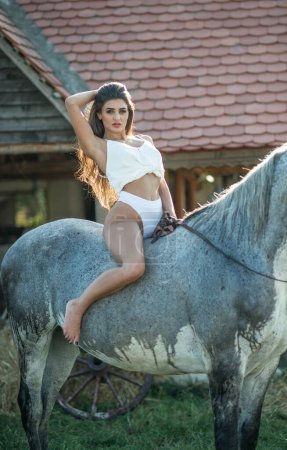 Photo for Beautiful brunette woman with long hair , white bikini and white shirt rides  on a white horse under a water drops. Sensual and attractive  girl  interacting and having fun with a horse at the ranch - Royalty Free Image