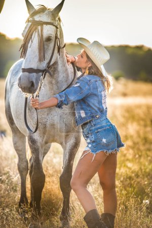 Foto de Hermosa mujer rubia con pelo rizado con sombrero blanco y caballo. Retrato de una chica con denim y su caballo. Hermosa chica interactuando y divirtiéndose con un caballo en el rancho - Imagen libre de derechos