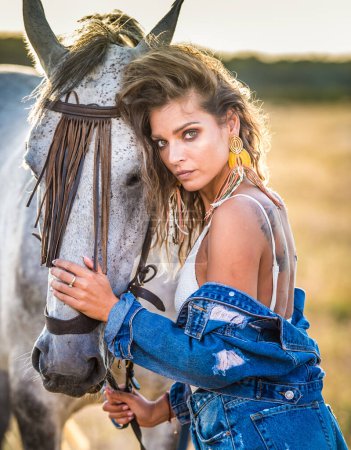 Photo for Beautiful blonde woman with curly hair with white hat and horse. Portrait of a girl with denim and her horse. Beautiful girl interacting and having fun with a horse at the ranch - Royalty Free Image