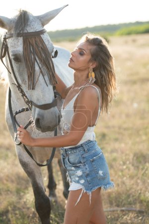 Photo for Beautiful blonde woman with curly hair with white hat and horse. Portrait of a girl with denim and her horse. Beautiful girl interacting and having fun with a horse at the ranch - Royalty Free Image
