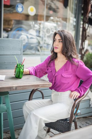 Photo for Happy Brunette Girl with pink blouse Sitting at the bar , Drinking a glass of lemonade While Smiling at the camera. Young sexy pretty woman on the chair  drinking  juice wearing a white pants - Royalty Free Image