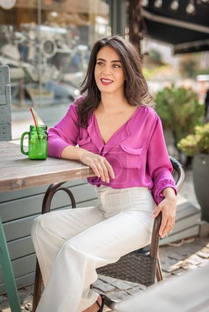 Photo for Happy Brunette Girl with pink blouse Sitting at the bar , Drinking a glass of lemonade While Smiling at the camera. Young sexy pretty woman on the chair  drinking  juice wearing a white pants - Royalty Free Image