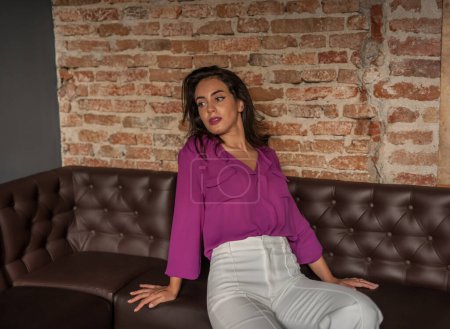 Téléchargez les photos : Belle jeune femme brune en chemisier violet et pantalon blanc posant à l'intérieur contre un mur de briques. Portrait d'une belle jeune femme brune - en image libre de droit