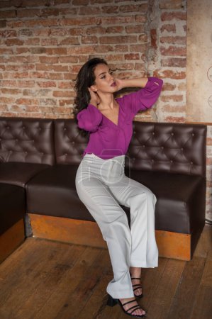 Foto de Hermosa mujer morena joven en una blusa púrpura y pantalones blancos posando en interiores contra una pared de ladrillo. Retrato de una hermosa joven morena - Imagen libre de derechos