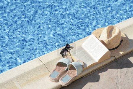 Stylish sunglasses, slippers, straw hat and book at poolside on sunny day, space for text. Beach accessories