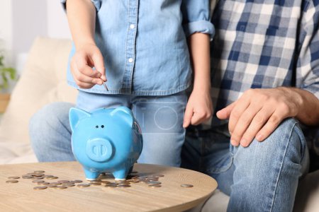 Little girl with her father putting coin into piggy bank at home, closeup