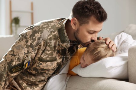 Foto de Soldado en uniforme militar ucraniano besando a su hija mientras dormía en el sofá en casa. Reunión familiar - Imagen libre de derechos