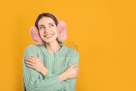 Happy woman wearing warm earmuffs on yellow background, space for text