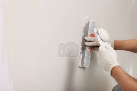 Worker plastering wall with putty knife indoors, closeup. Space for text