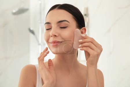Schöne junge Frau tut Gesichtsmassage mit gua sha Werkzeug im Badezimmer