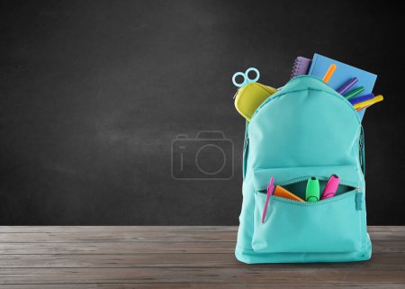 Backpack with school stationery on wooden table near black chalkboard, space for text