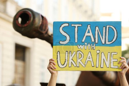 Frau hält Plakat in den Farben der Nationalflagge mit den Worten "Stand with Ukraine near broken tank on city street", Nahaufnahme