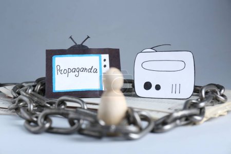 Concepto de propaganda. Humanos atrapados en el campo de los medios. Figura de madera encadenada, periódicos, televisión de papel y radio en la mesa