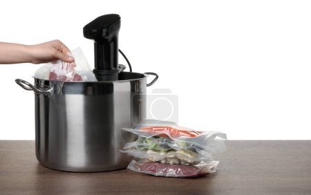 Woman putting vacuum packed meat into pot with sous vide cooker on wooden table against white background, closeup. Thermal immersion circulator