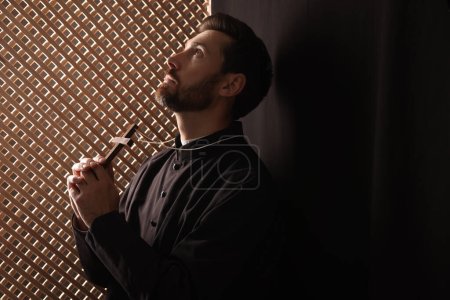 Katholischer Priester in Soutane mit Kreuz und Gebet zu Gott im Beichtstuhl, Raum für Text