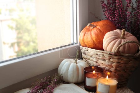 Beautiful heather flowers, burning candles, open book and wicker basket with pumpkins near window indoors. Space for text