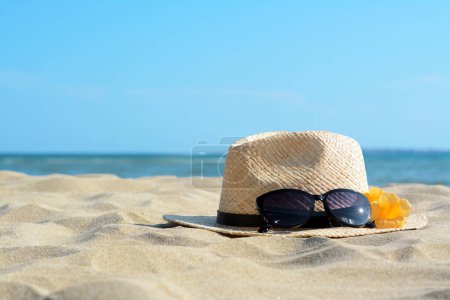 Téléchargez les photos : Chapeau avec de belles lunettes de soleil et des fleurs sur le sable près de la mer. Espace pour le texte - en image libre de droit