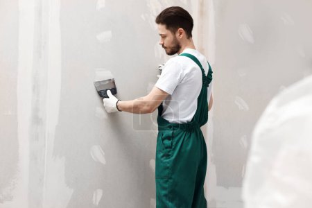 Photo for Worker in uniform plastering wall with putty knife indoors - Royalty Free Image