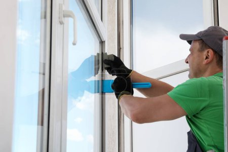 Téléchargez les photos : Travailleur en uniforme utilisant une spatule pour l'installation de fenêtres à l'intérieur - en image libre de droit