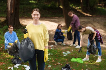 Téléchargez les photos : Jeune femme avec sac en plastique et groupe de personnes ramassant des ordures dans le parc - en image libre de droit
