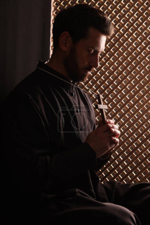 Sacerdote católico en sotana sosteniendo cruz en cabina confesional