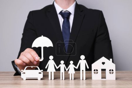 Man holding figure of umbrella near family, house and car at wooden table. closeup. Insurance concept