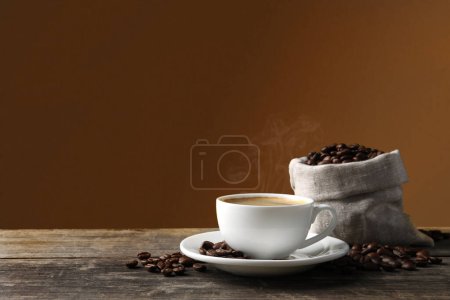 Photo for Cup of hot aromatic coffee and roasted beans on wooden table against brown background. Space for text - Royalty Free Image