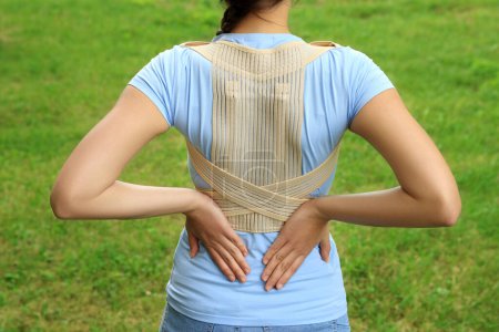 Closeup of woman with orthopedic corset on green grass outdoors, back view
