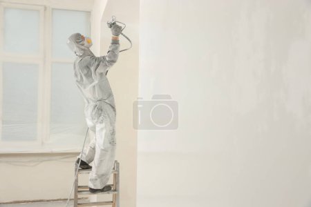 Téléchargez les photos : Décorateur en salopette de protection peinture murale avec pistolet de pulvérisation à l'intérieur - en image libre de droit