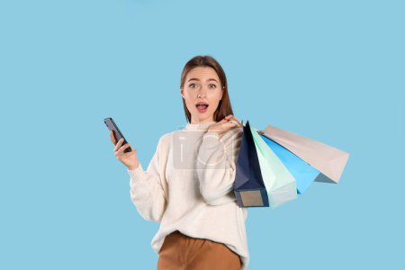 Téléchargez les photos : Surprise jeune femme avec des sacs à provisions et smartphone sur fond bleu clair. Grande vente - en image libre de droit