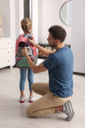 Father putting bottle of water into daughter`s backpack at home. Preparing to school