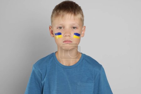 Kleiner Junge mit Zeichnung der ukrainischen Flagge im Gesicht vor hellgrauem Hintergrund