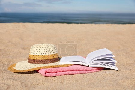 Foto de Libro abierto, sombrero y toalla rosa en la playa de arena cerca del mar. Espacio para texto - Imagen libre de derechos