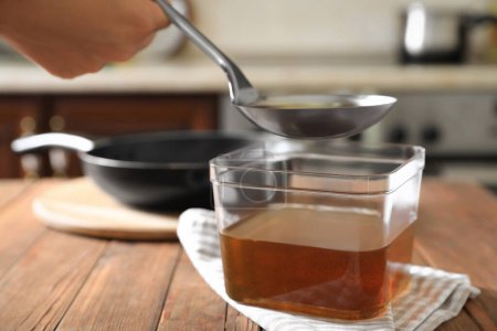 Woman pouring used cooking oil with ladle into container on wooden table in kitchen, closeup