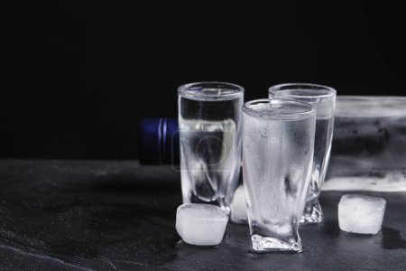 Foto de Botella de vodka y vasos de chupito con hielo sobre la mesa sobre fondo negro. Espacio para texto - Imagen libre de derechos