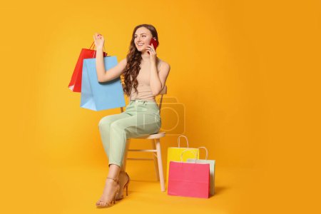 Photo for Beautiful woman holding colorful shopping bags and talking on smartphone against orange background - Royalty Free Image