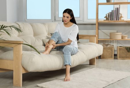 Young woman rubbing sore leg on sofa at home Poster 625062090